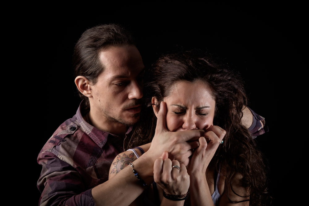 A man in a striped shirt grips a woman's face aggressively, covering her mouth. She appears distressed, eyes tightly closed, set against a black backdrop.