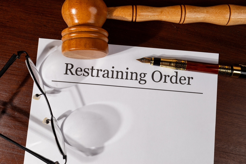 A judge's gavel and glasses rest on a "Restraining Order" document placed on a wooden desk. The tone is serious, related to legal matters.