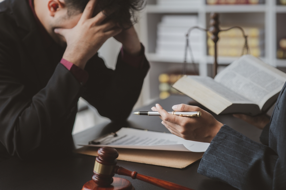 Stressed individual consulting with an attorney at a law office, with legal documents, a gavel, and scales of justice visible on the desk.