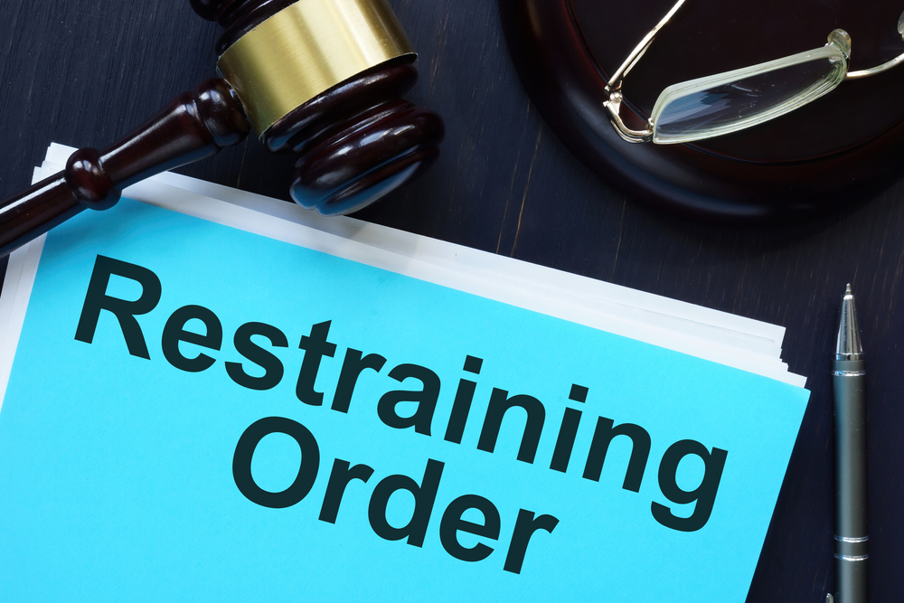 A blue document labeled "Restraining Order" lies on a dark table beside a judge's gavel, a pair of glasses, and a pen, conveying a legal and serious tone.
